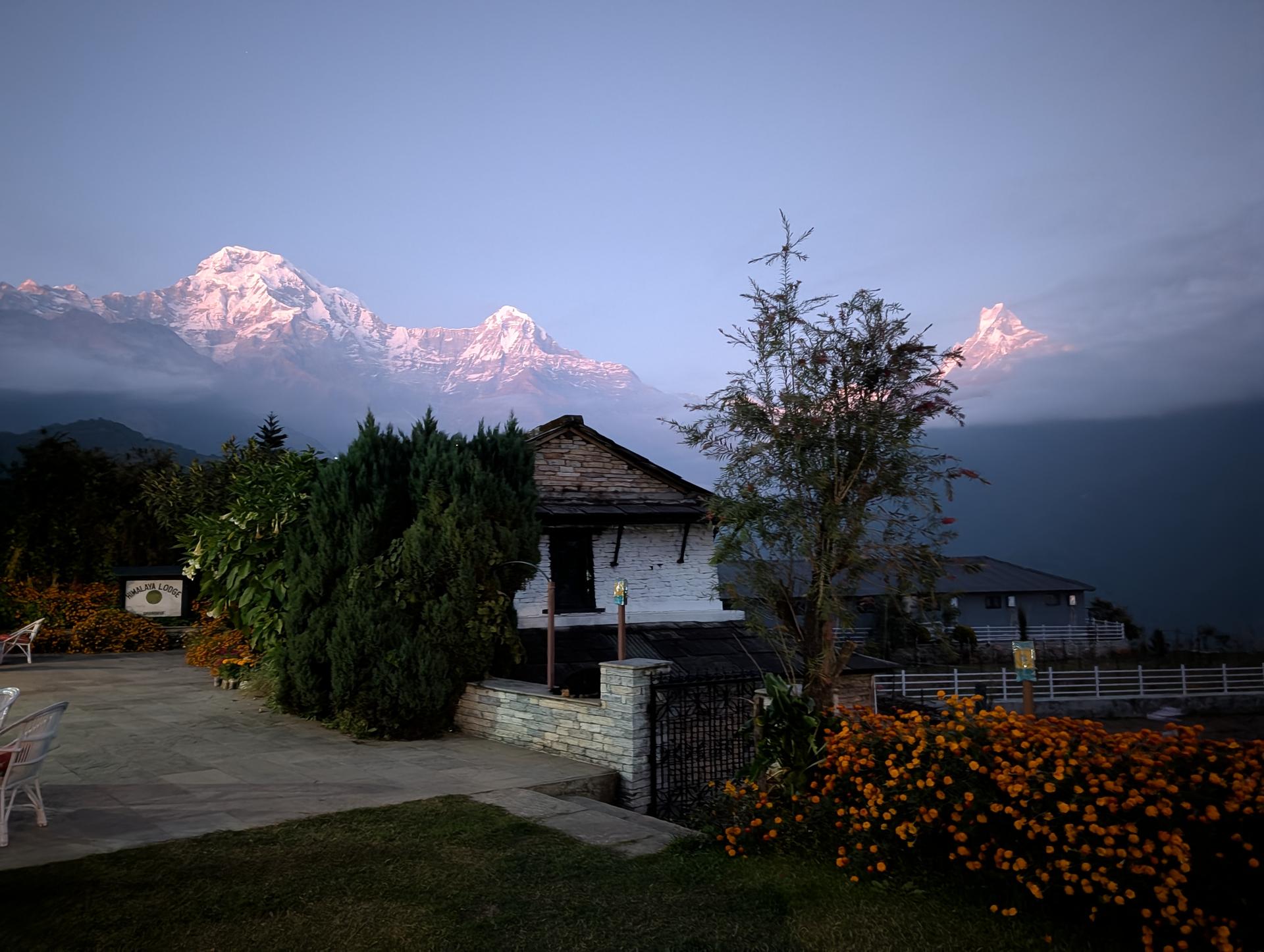 A small lodge and garden sit beneath pink-tinted snow-capped peaks at dawn, with clouds and mist drifting across the mountains.