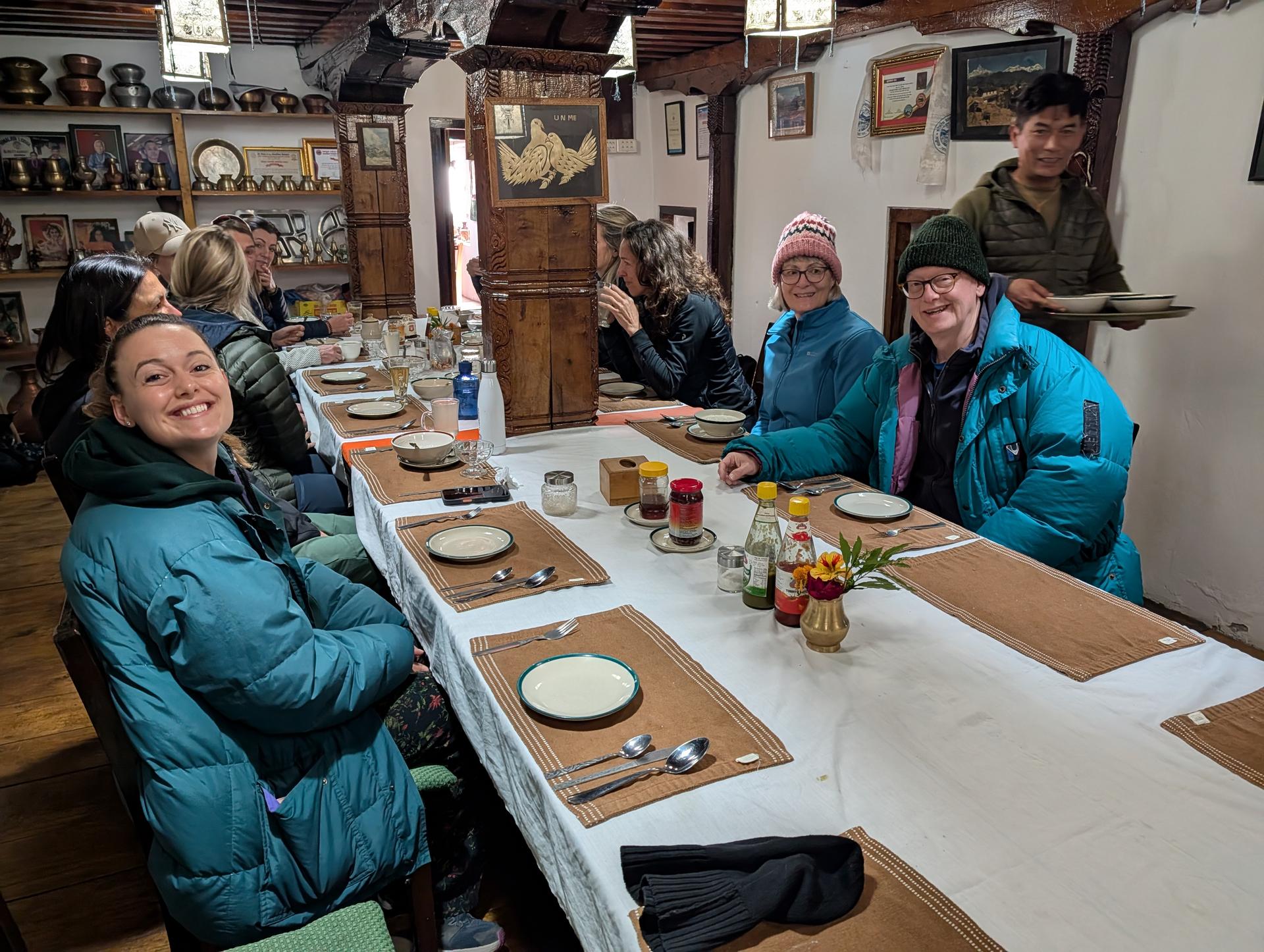 A long dinner table inside a cozy lodge, set with plates and condiments, as a group chats while a server carries dishes.