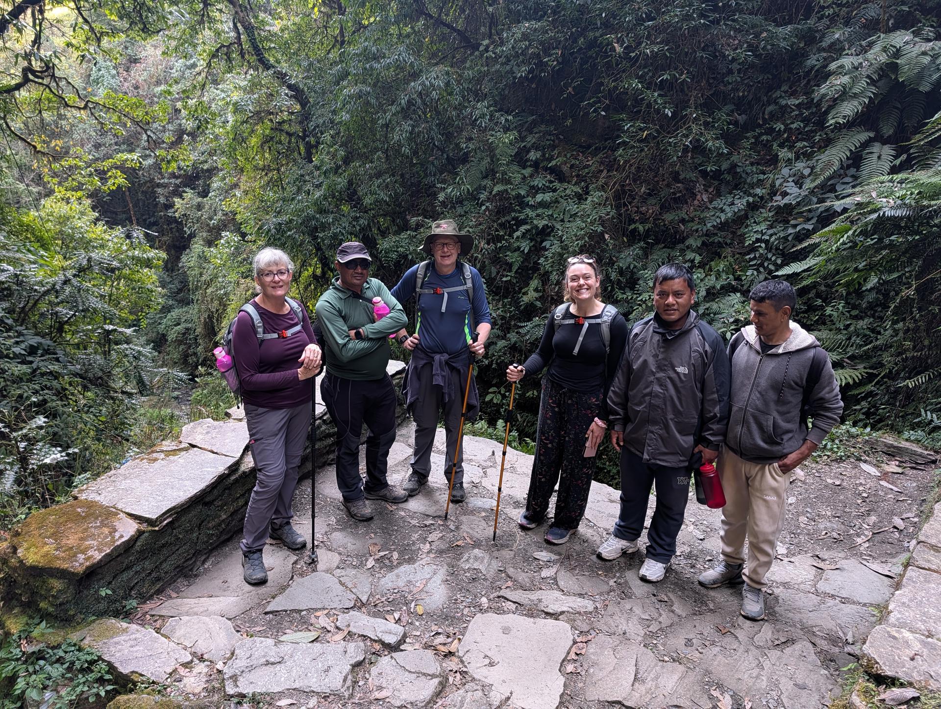 A group of hikers pose on a stone path in a lush forest ravine, with trekking poles and water bottles.