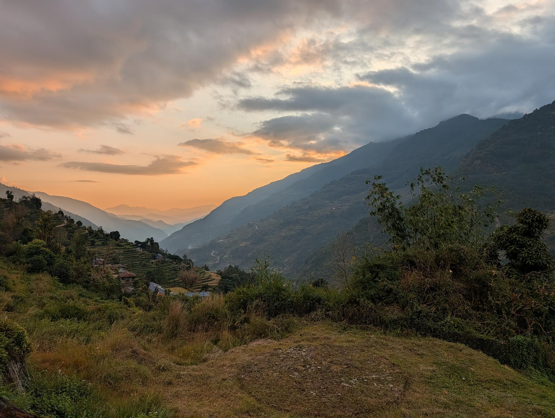 Sunset over layered mountain ridges, with orange light on clouds and terraced hillsides in the foreground.