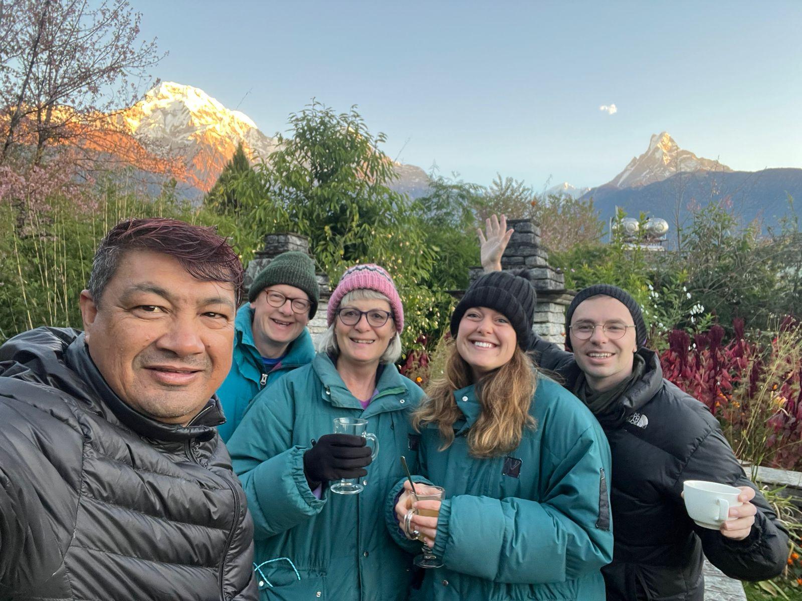 A group selfie of five smiling travelers in warm jackets, holding hot drinks, with misty mountains and a pointed peak in the background.