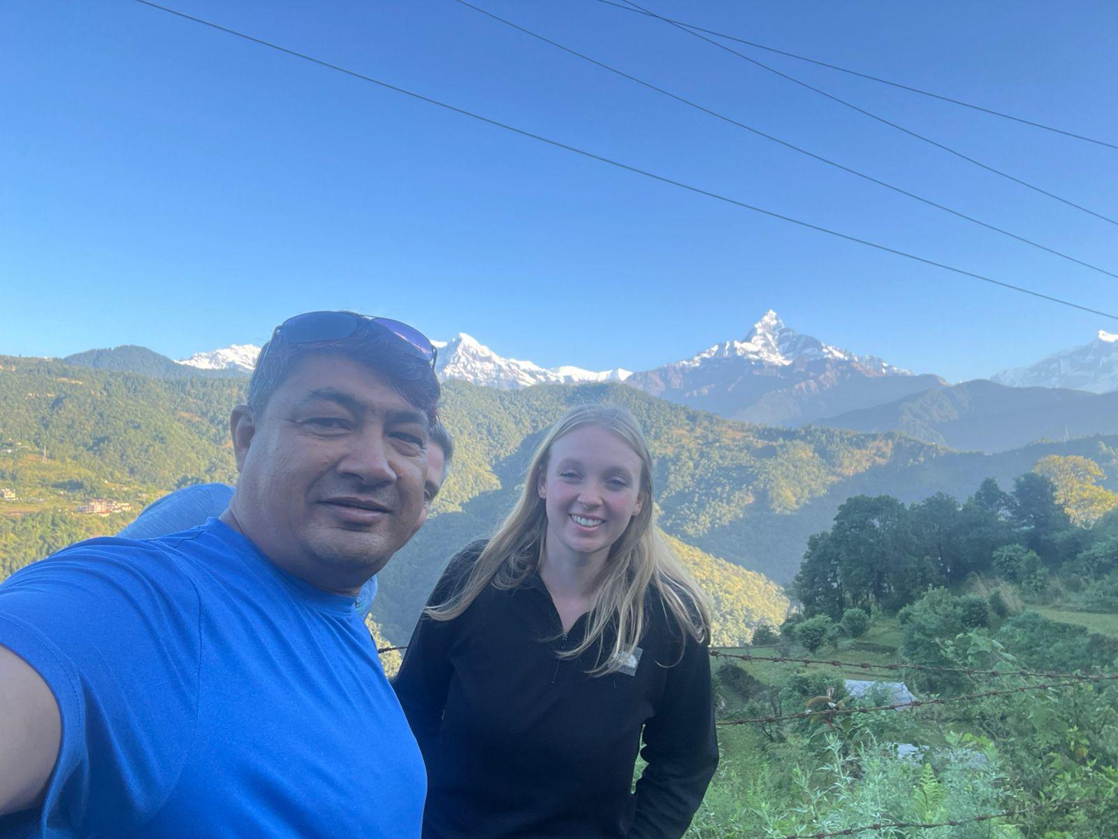 Two people take a selfie at a scenic overlook, with green hills and a sharp Himalayan peak under a bright blue sky.