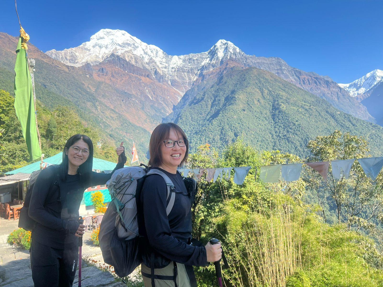 Two hikers with backpacks and trekking poles stand near a lodge, smiling with terraced hills and snow-capped mountains behind them.