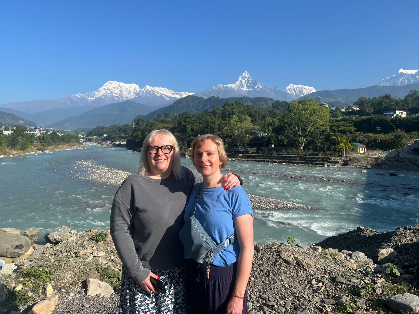 Two women stand by a wide river with mountains and a sharp snowy peak in the distance on a clear sunny day.