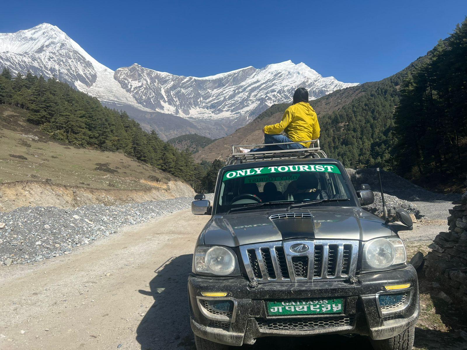 A traveler in a yellow jacket sits on the roof of a dusty SUV on a mountain road, facing dramatic snow-covered peaks.