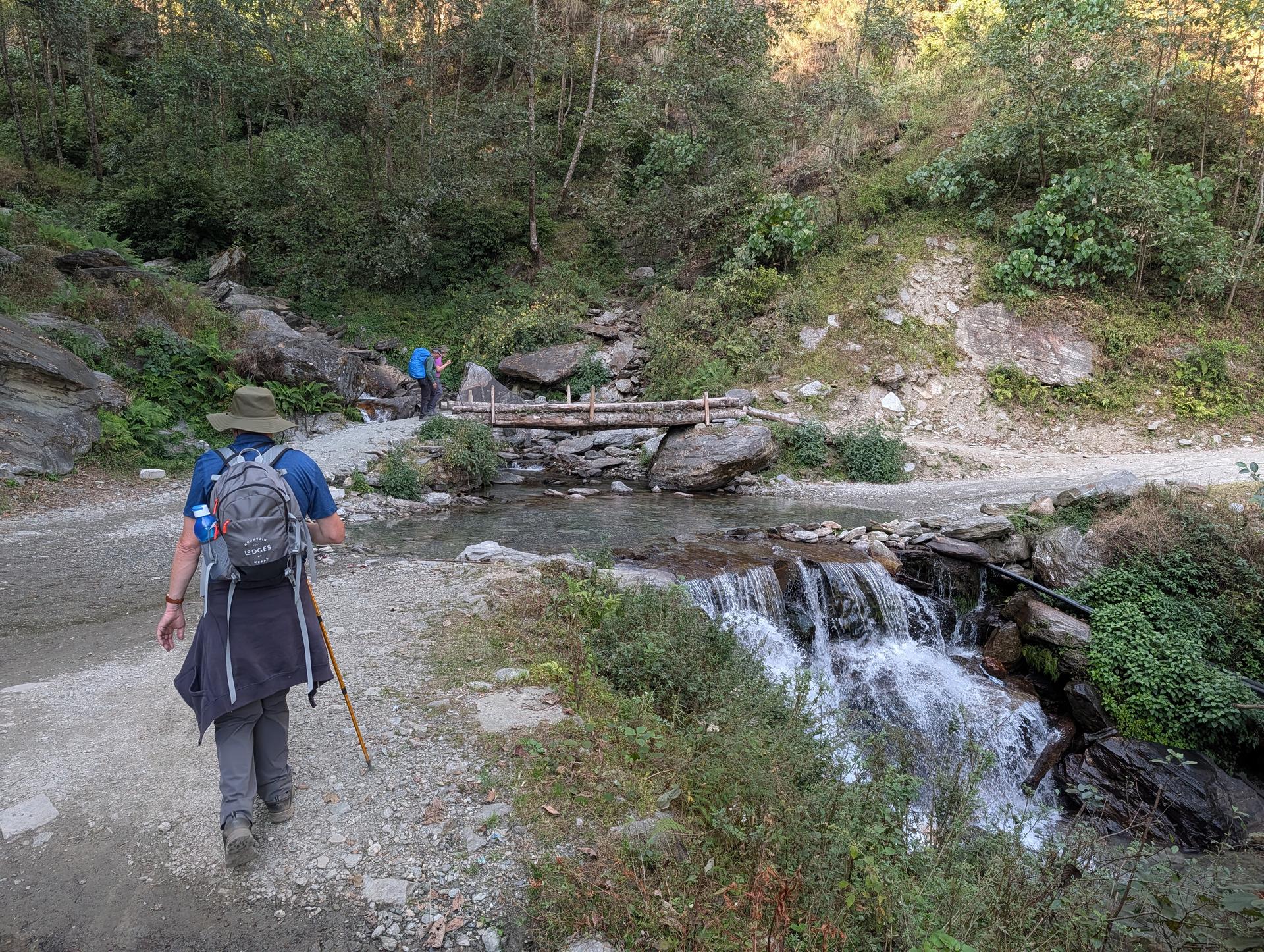 A hiker with a backpack approaches a small wooden bridge over a stream, with a short waterfall and rocky forested hillside nearby.