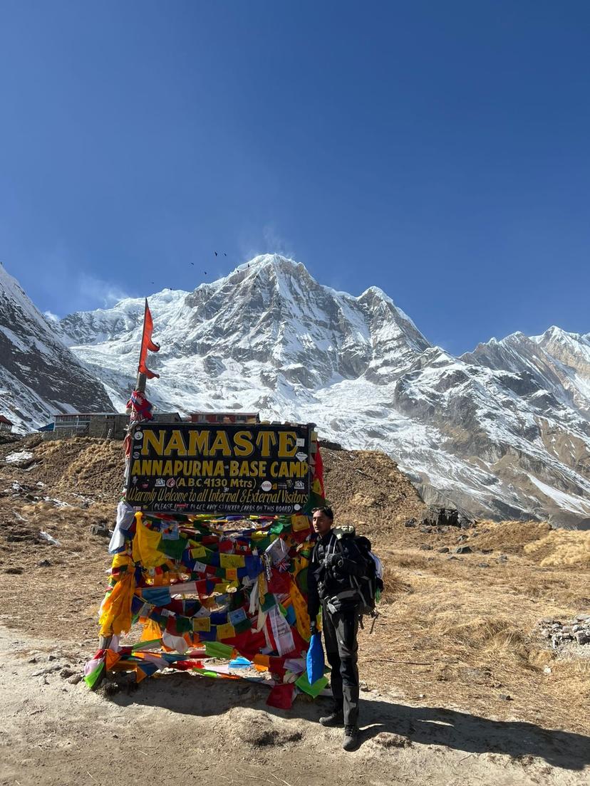 A tourist in high altitude hiking gear stands in front of a welcome sign for Annapurna Base Camp which is covered with colourful prayer flags.