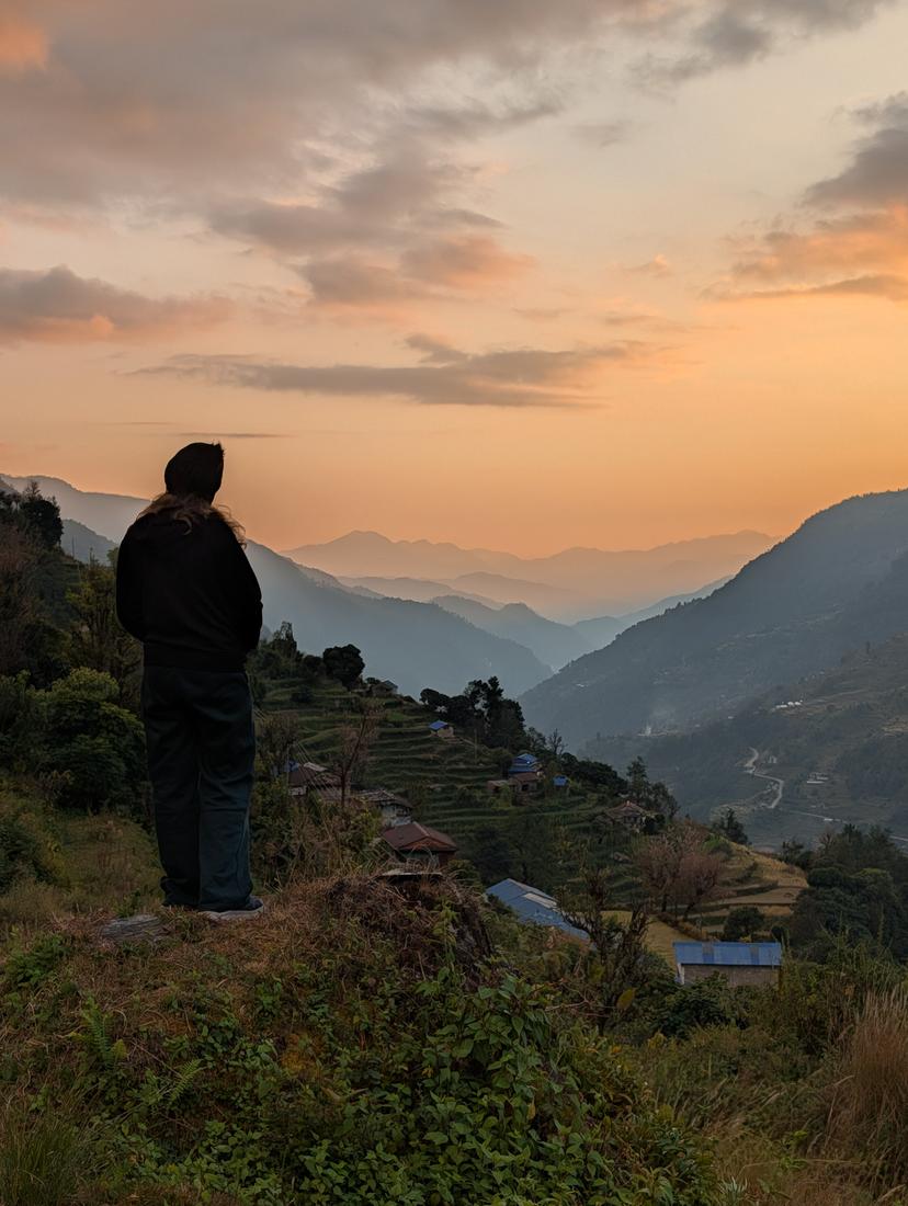 A tourist in winter clothing stands sillouhetted against a golden sky overlooking the Pokhara Valley